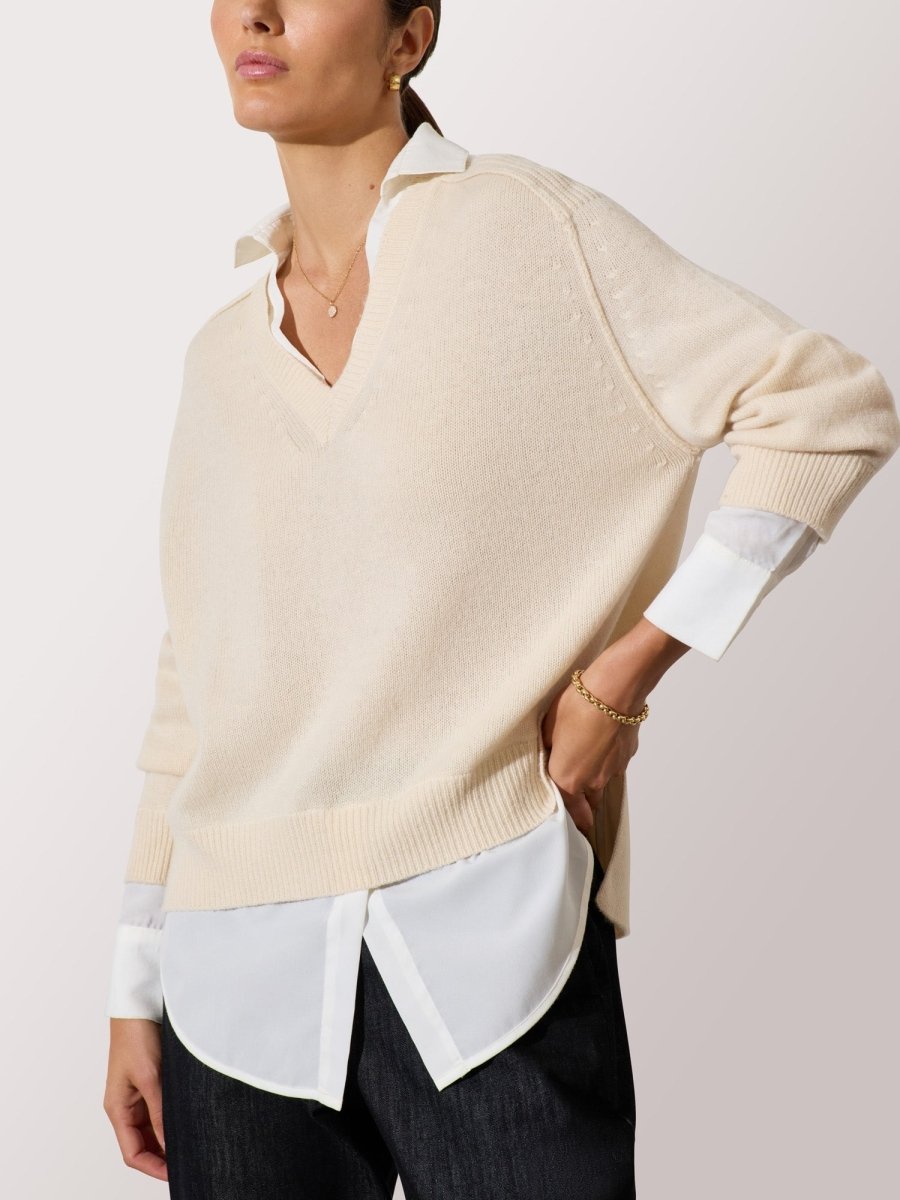 Women's cardigan sweater in light beige layered over a white blouse and paired with dark denim trousers.