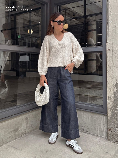 Woman wearing wide-leg denim pants with a textured white knit sweater, accessorized with sneakers and a woven handbag for a relaxed city look.